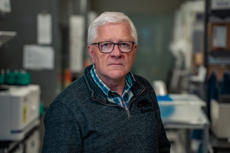 John Dick, a man with white hair stands inside a lab, with a serious look on his face.